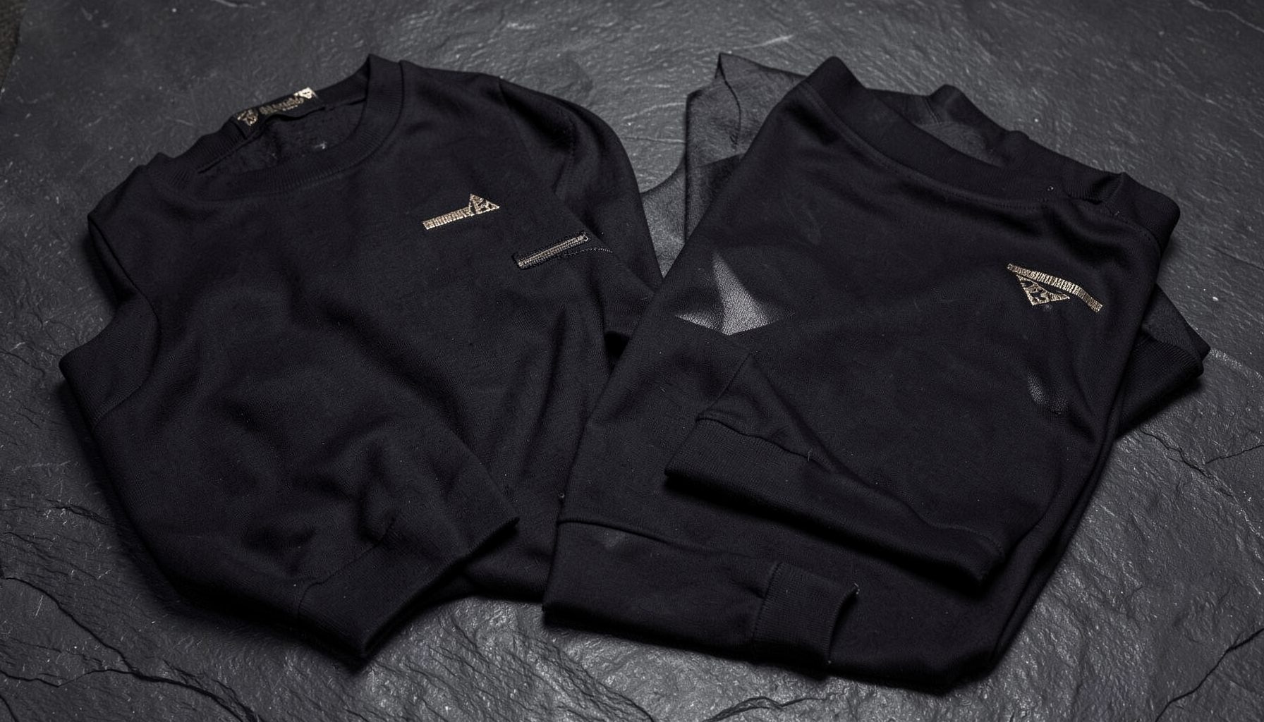 Dark, moody apparel pieces with subtle metallic accents, arranged on a textured slate background.